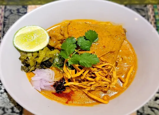 Khao Soi (Northern Thai Curry Noodles) — Egg Noodles, chicken or beef in the coconut curry soup, top with pickled mustard greens, shallots, crispy noodles, chili oil, and lime.